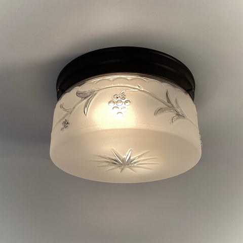 Illuminated 1940s frosted glass flush-mount ceiling light with embossed grapevine and starburst design on dark bronze canopy.