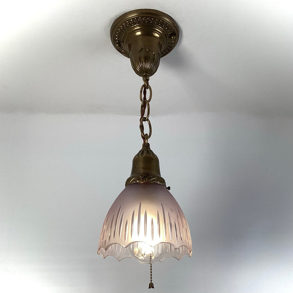 1920s antique brass pendant light with frosted fluted glass shade and pull-chain, illuminated and hanging from ornate ceiling canopy.